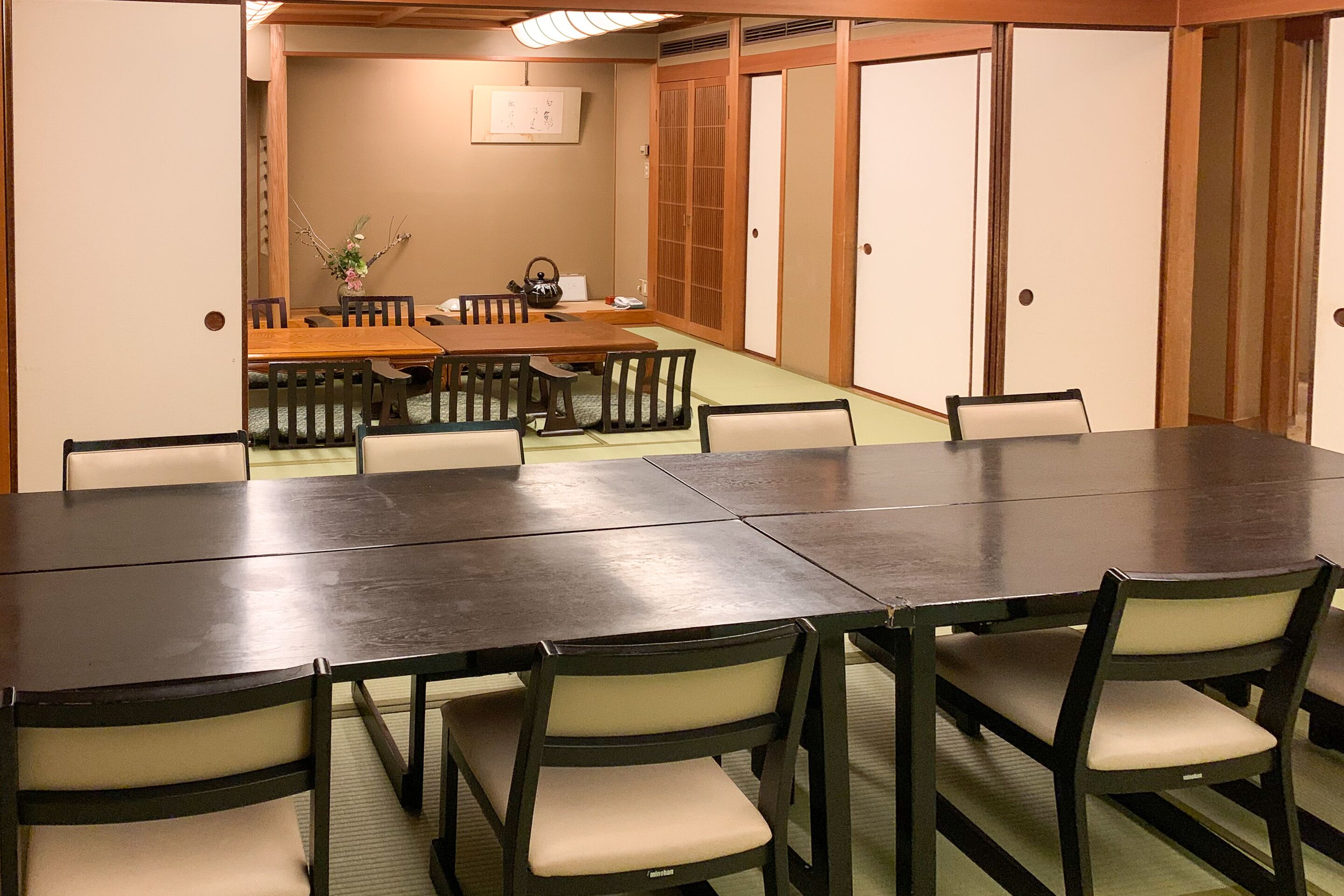 Una sala conferenze con un lungo tavolo e sedie, porte scorrevoli in legno e un tavolino con un bollitore e fiori.
