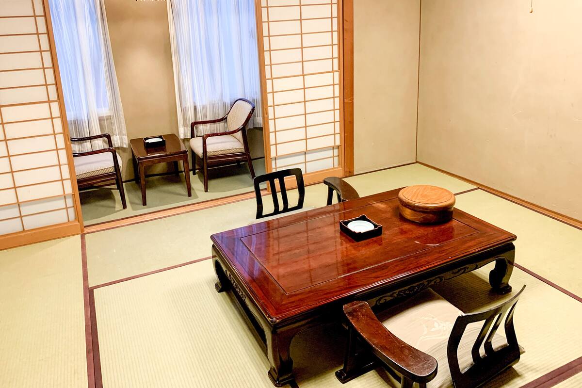 A traditional Japanese room with tatami flooring, a wooden table with a cushion, and two chairs.