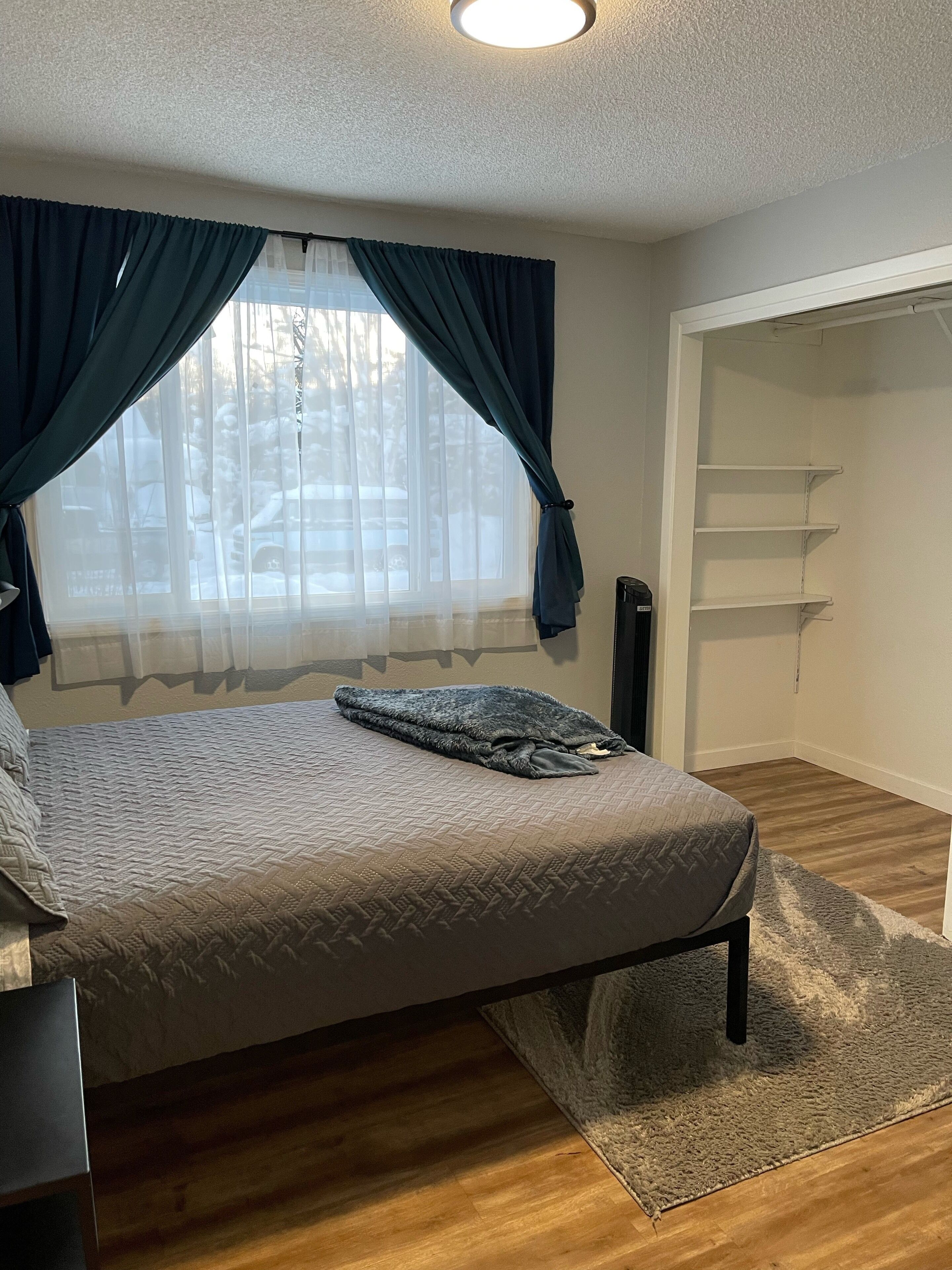 Une chambre à coucher avec un lit, une fenêtre avec des rideaux, un plancher en bois et une armoire avec des étagères.