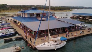 Un restaurante frente al agua con terraza, un velero amarrado cerca y vista al agua y la costa distante.