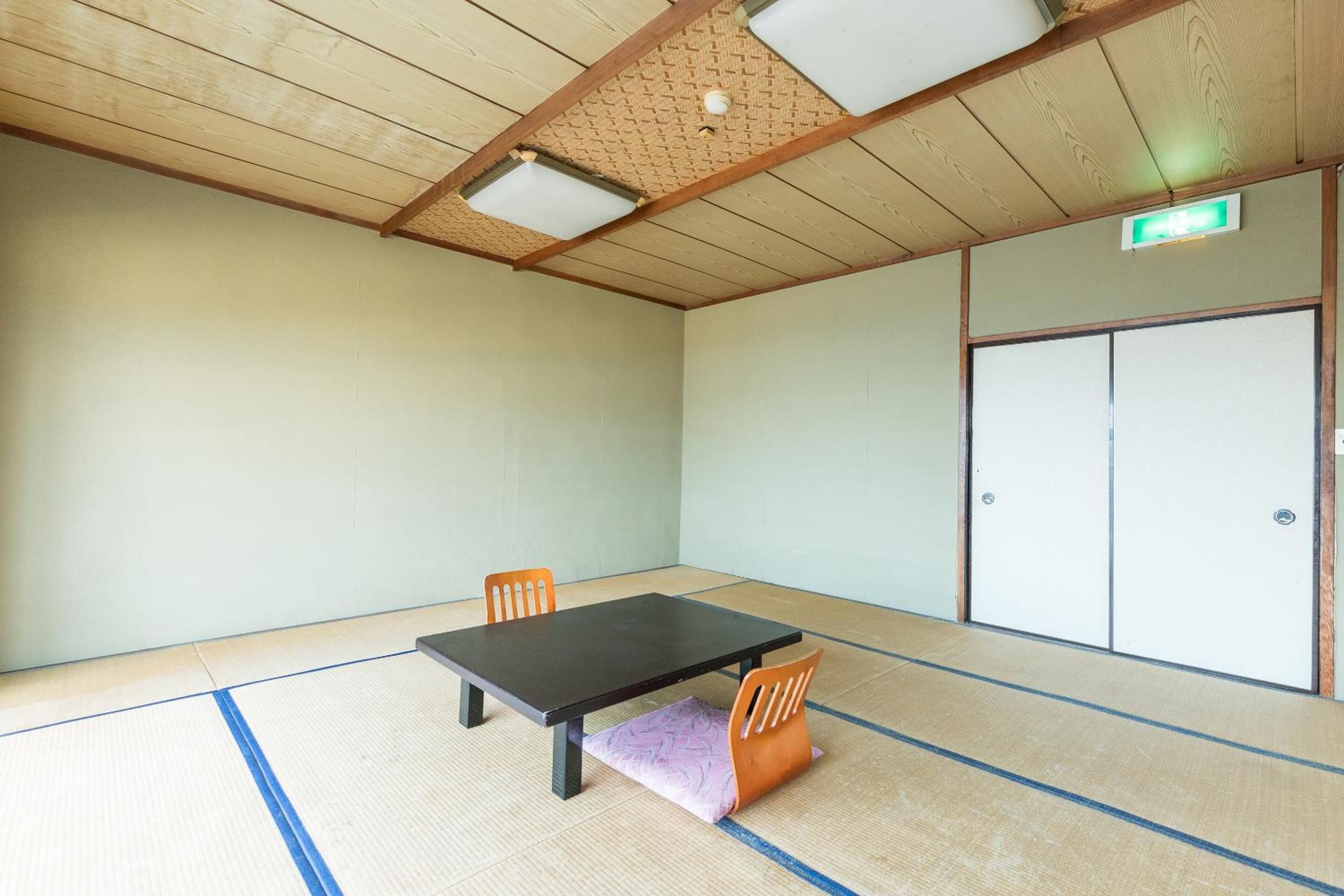 A room with tatami flooring, a black table, two orange chairs, and a wooden ceiling with recessed lighting.