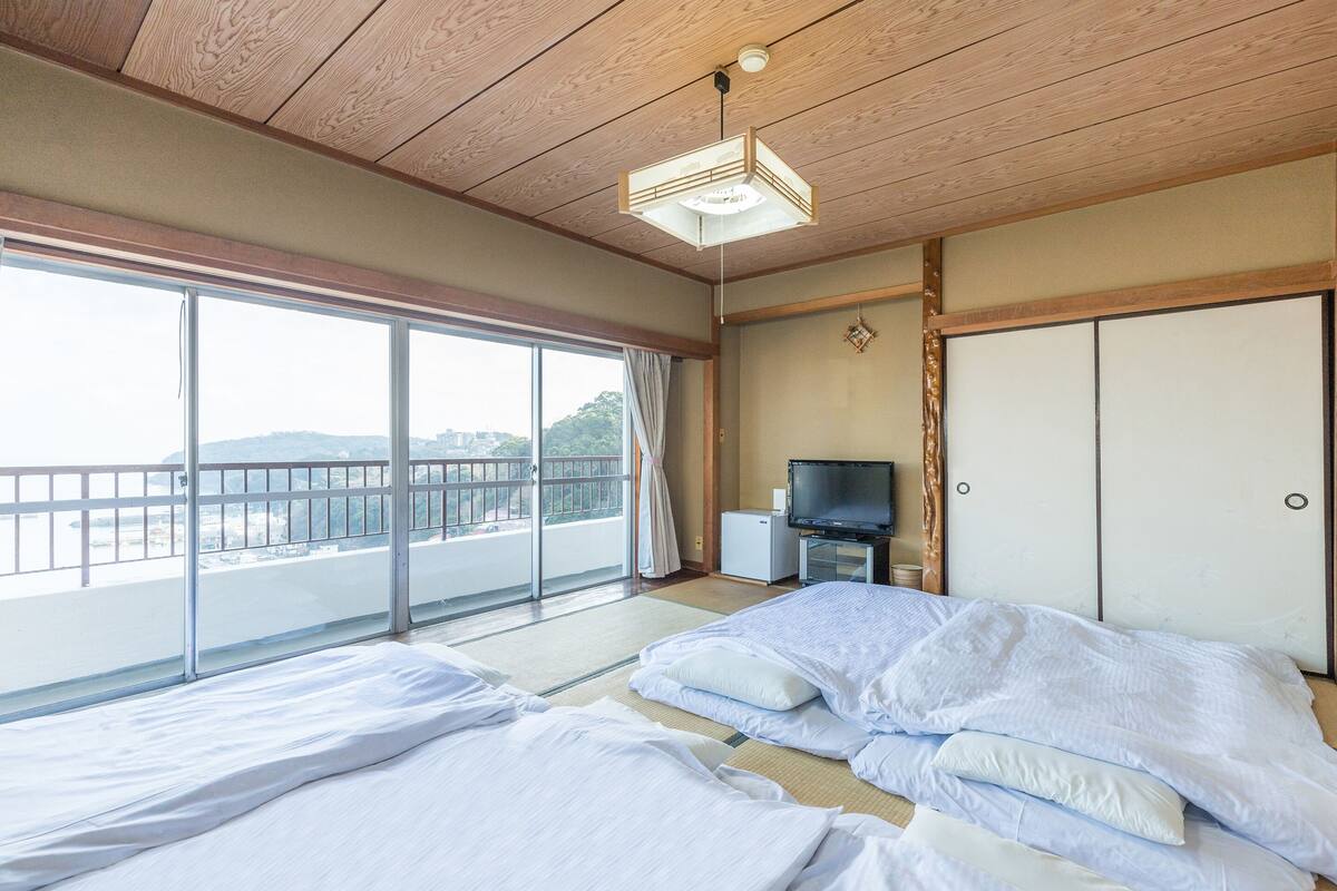 Ett hotellrum med en stor skjutdörr i glas som leder ut till en balkong, en säng med vita sängkläder, en liten tv och en traditionell japansk garderob.