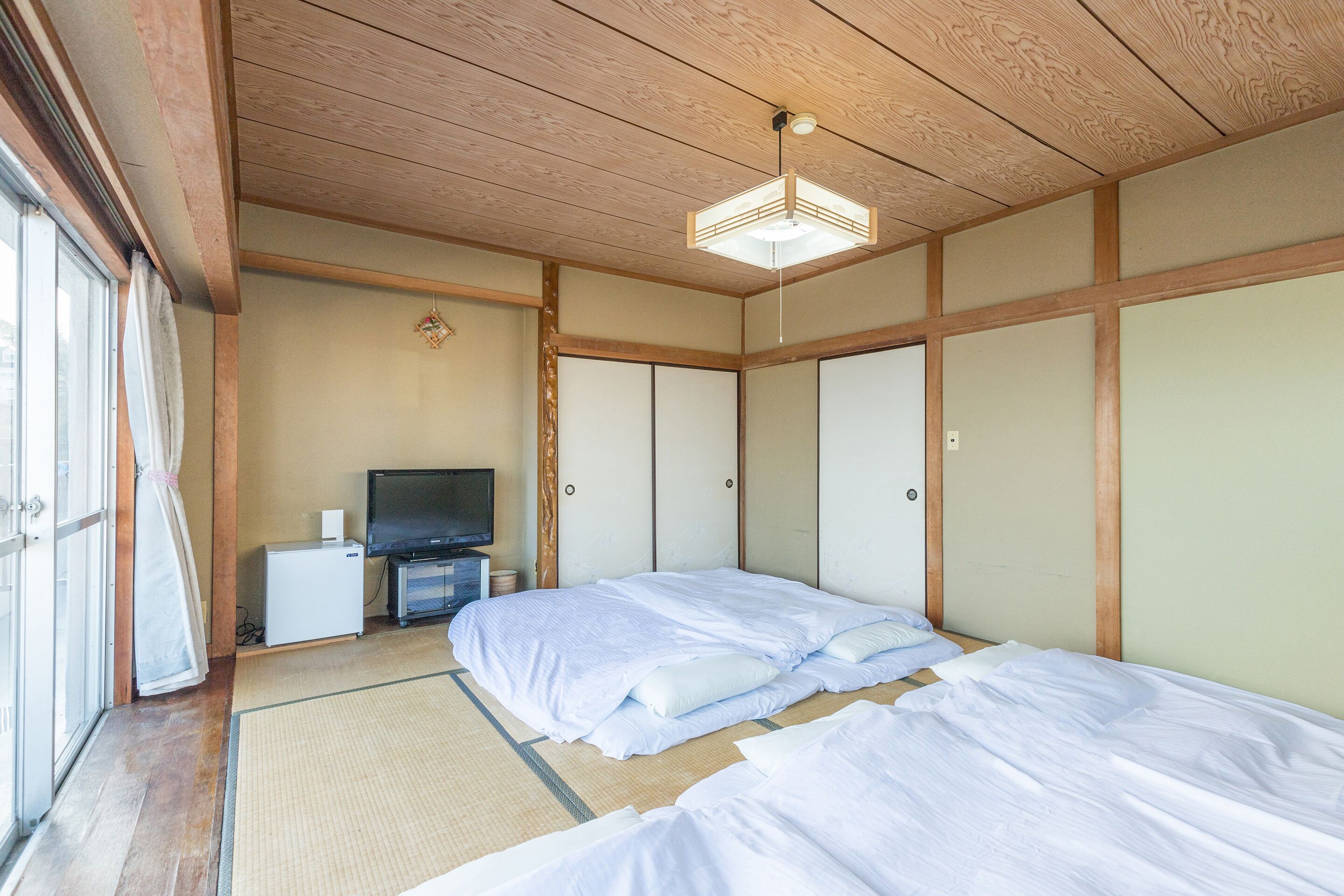 A traditional Japanese-style room with tatami flooring, a bed with white bedding, a television on a small table, and sliding doors.