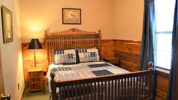 A wooden-framed bed with a plaid bedspread and pillows, a bedside table with a lamp, and a framed picture on the wall.