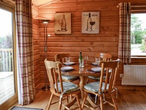A wooden cabin interior with a dining table set for four, wooden chairs with cushioned seats, a floor lamp, and two framed pictures on the wall.