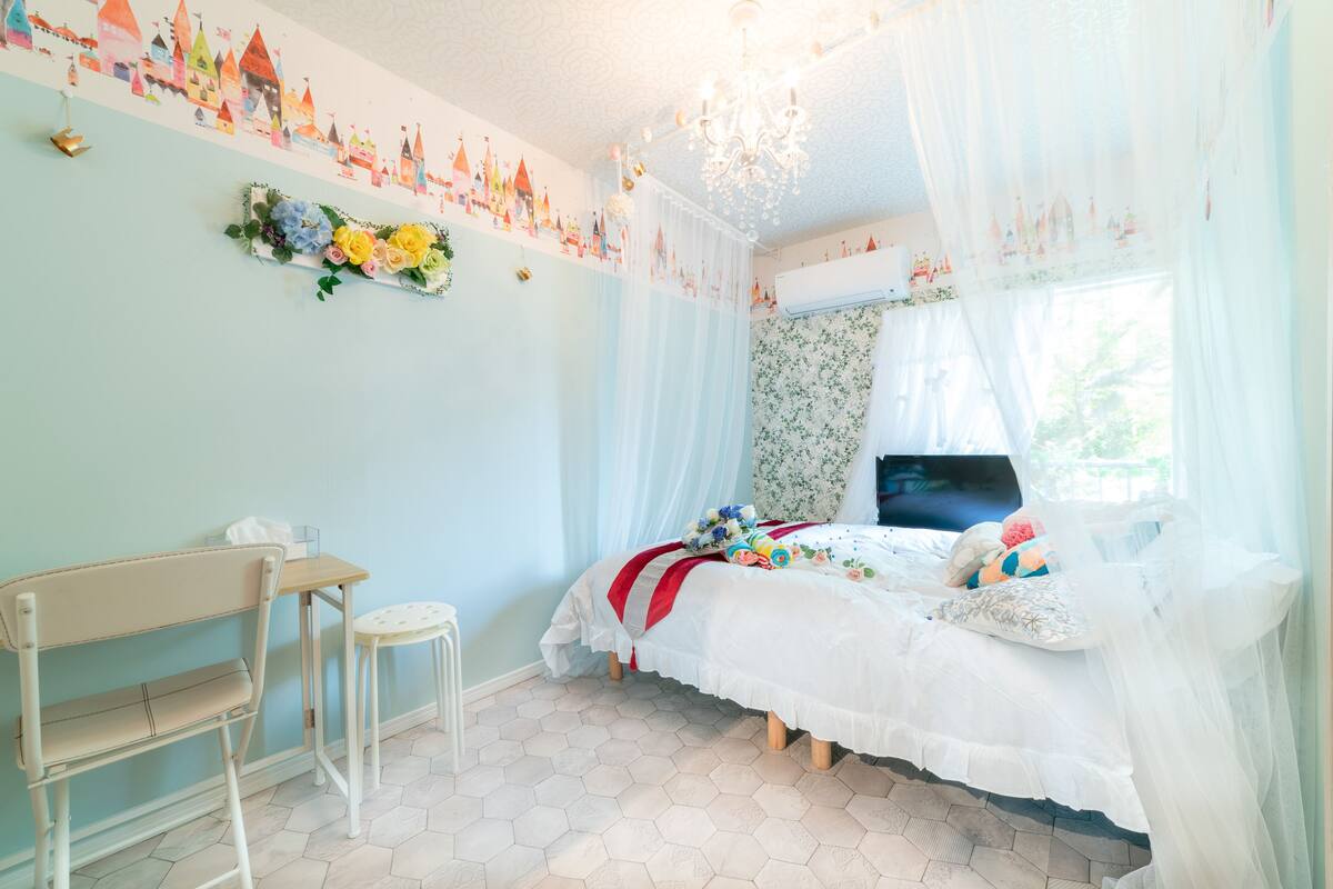 A bedroom with a bed, a desk, a chair, a television, and a window with sheer curtains.