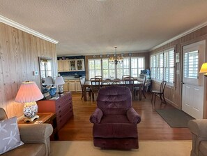 A spacious living area with a large armchair, wooden paneling, a dining table with chairs, and a kitchen in the background.