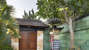 A traditional house with a tiled roof, a wooden door, and a small garden area with plants and a checkered cloth on a stand. - Villa Sotra Description (Denpasar)
