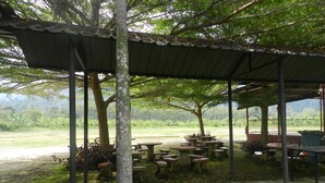 An outdoor patio area with tables and benches under a covered structure, surrounded by trees and a view of a green landscape. - The Red Cabanon (Janda Baik)