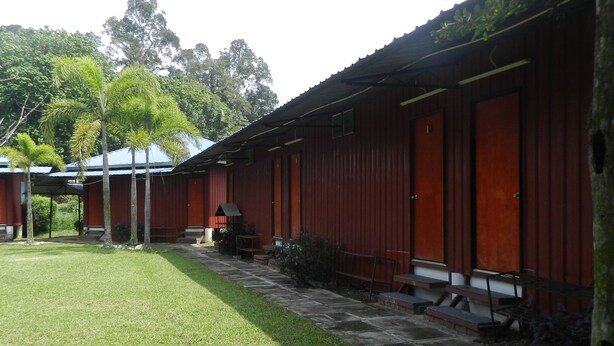 A row of red buildings with a grassy courtyard and trees in the background. - The Red Cabanon (Janda Baik)