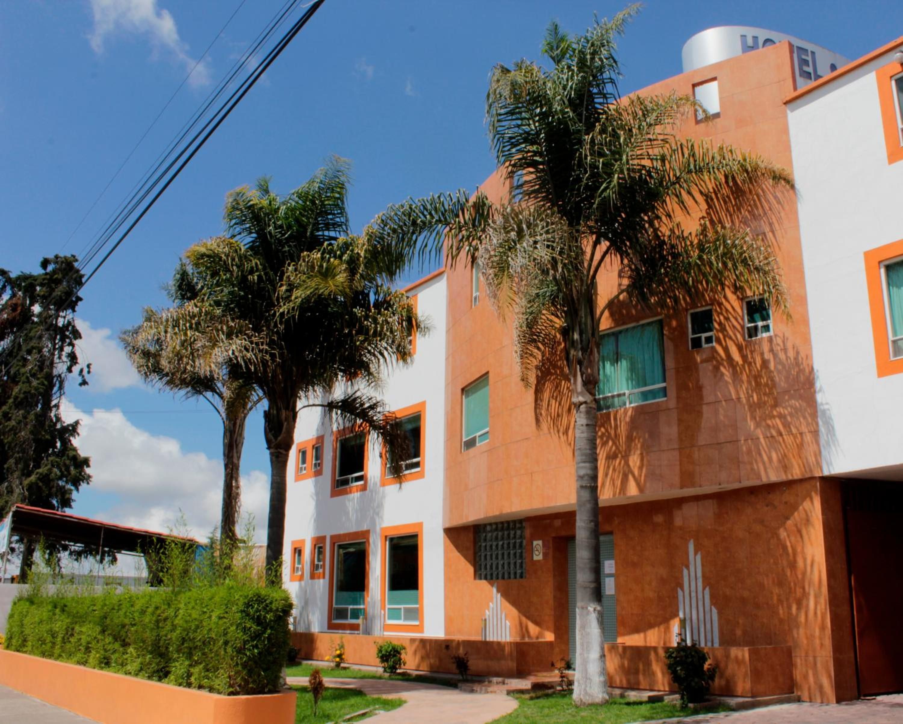 A modern building with multiple windows and a flat roof, surrounded by greenery and palm trees.