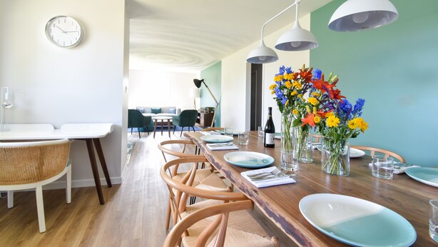 Un coin repas avec une longue table en bois, des chaises blanches et une horloge au mur.