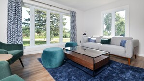 Un salon moderne avec un canapé, un pouf et une table basse, de grandes fenêtres avec des rideaux et une vue sur la verdure à l’extérieur.