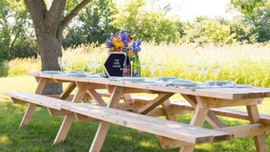 Une table de pique-nique en bois est installée à l’extérieur, avec une pancarte indiquant « The Vine House » et un vase rempli de fleurs.