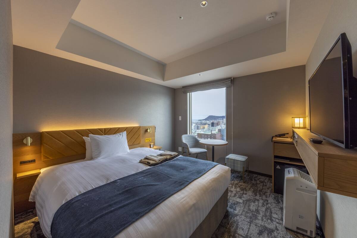 Habitación de hotel con una cama grande, un escritorio, una silla, un televisor y una ventana con vistas a la ciudad.