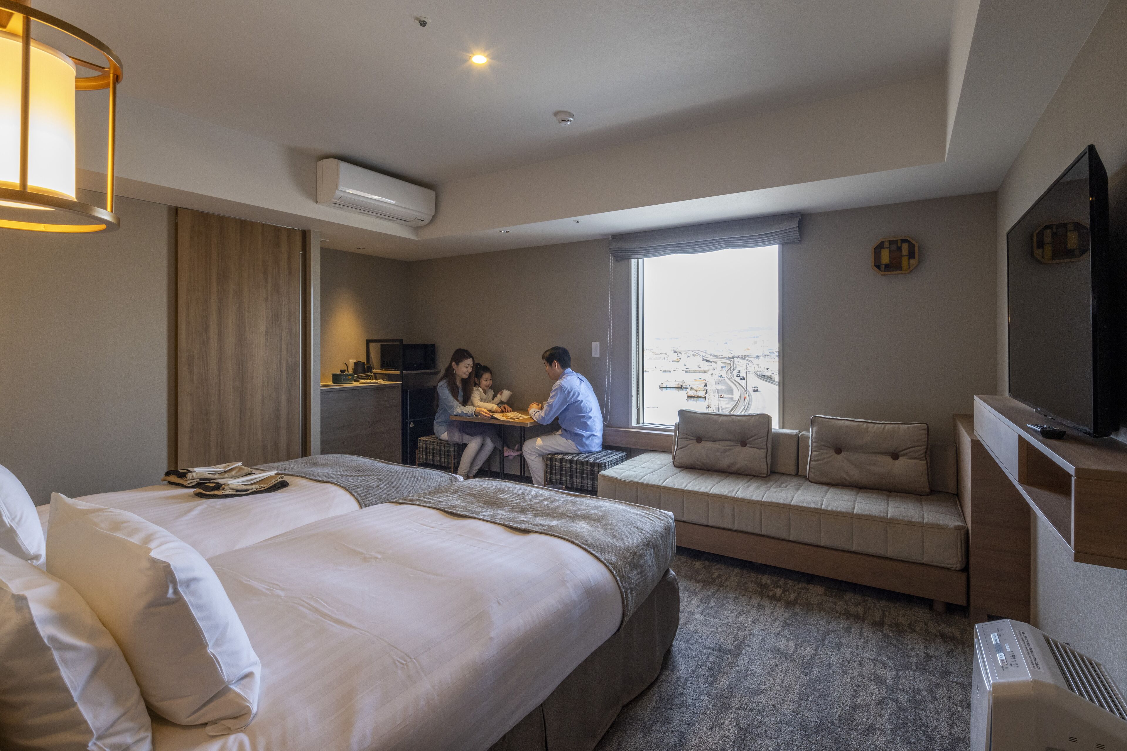 Une chambre d’hôtel avec un grand lit, un canapé-lit, un bureau où deux personnes travaillent, une télévision et une fenêtre donnant sur la ville.