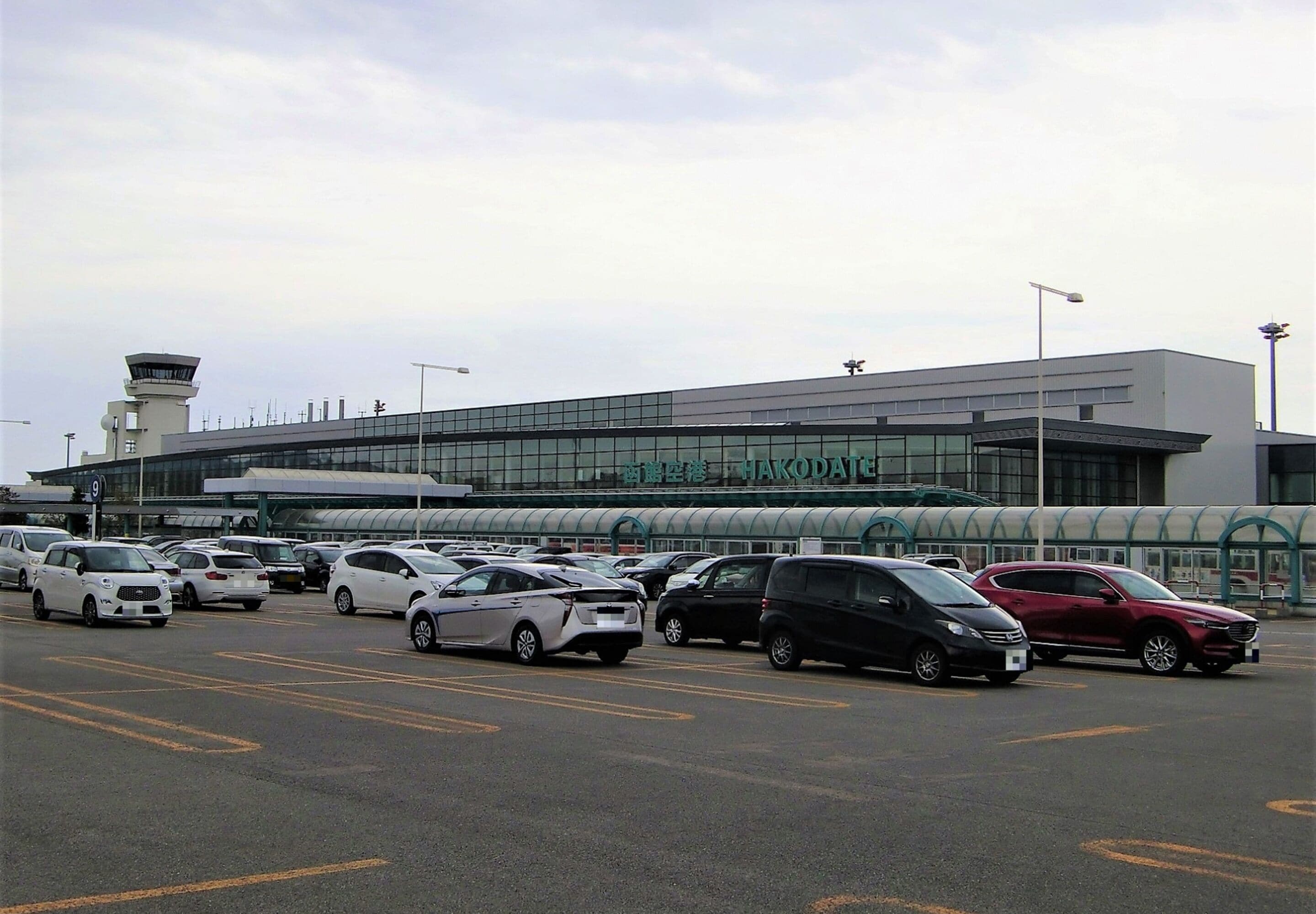 A parking lot with various cars and a modern building with a sign that reads "HAKODATE".