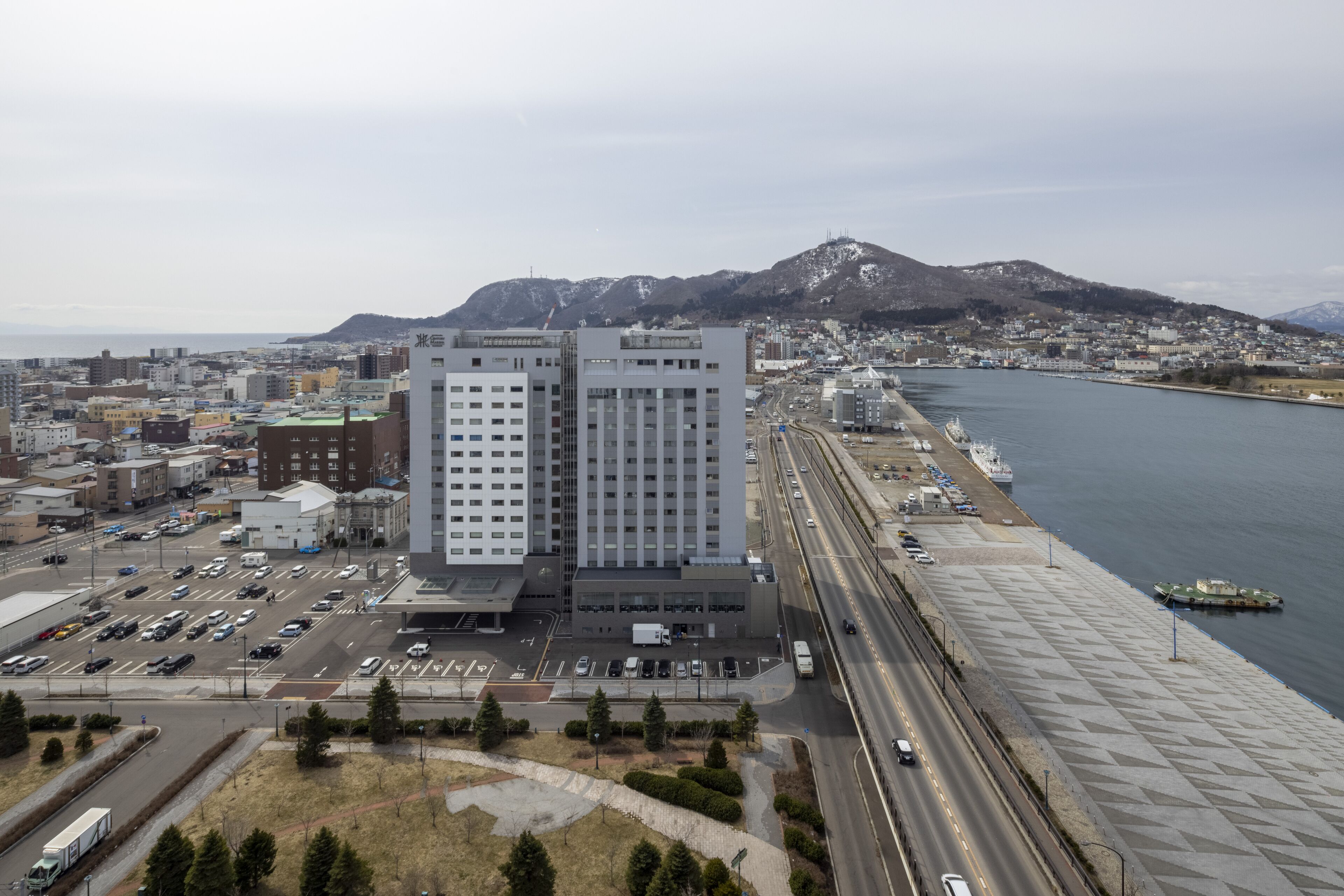 A high-rise building with a modern design, located near a waterfront with a road and parking area in the foreground.