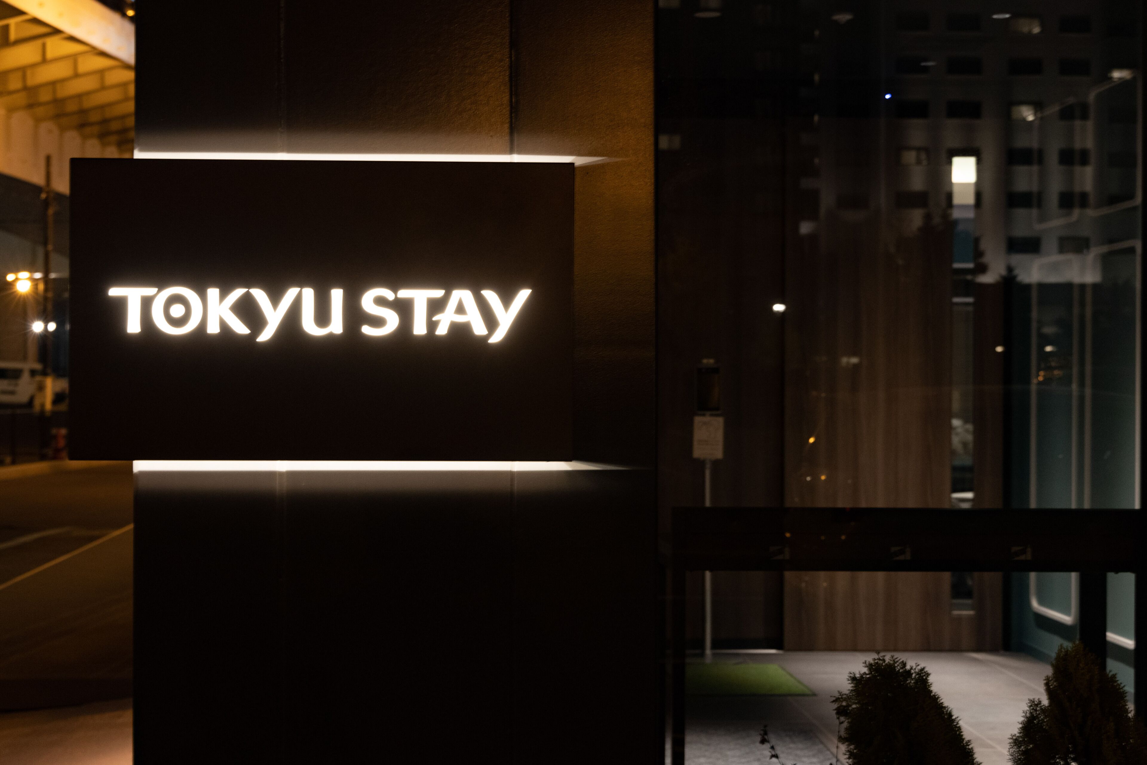 A sign for "TOKYU STAY" with a modern design, located at night with visible city lights in the background.