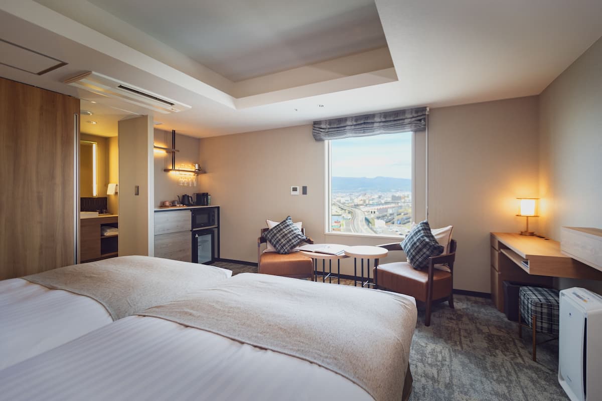 Habitación de hotel con una cama grande, un escritorio, una silla, una mesita y vistas a un paisaje urbano a través de un ventanal amplio.