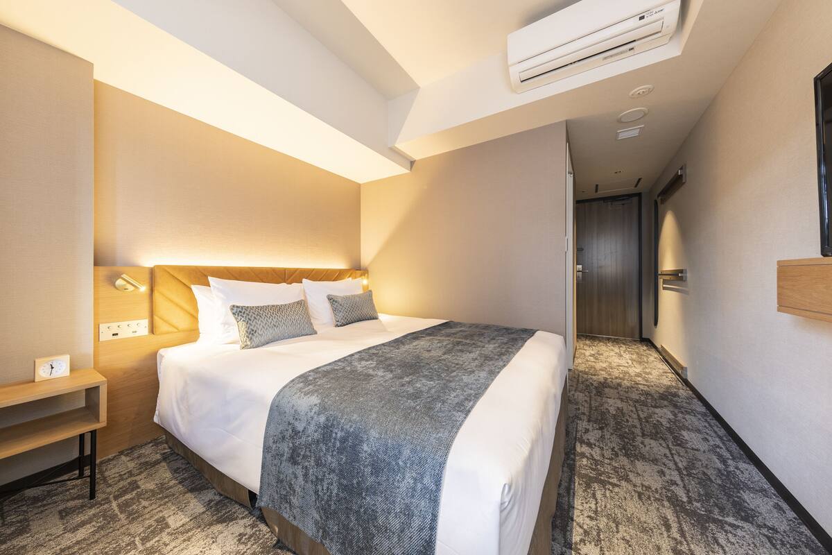 Una habitación de hotel con una cama grande, una mesita de noche, un equipo de aire acondicionado y suelo alfombrado.