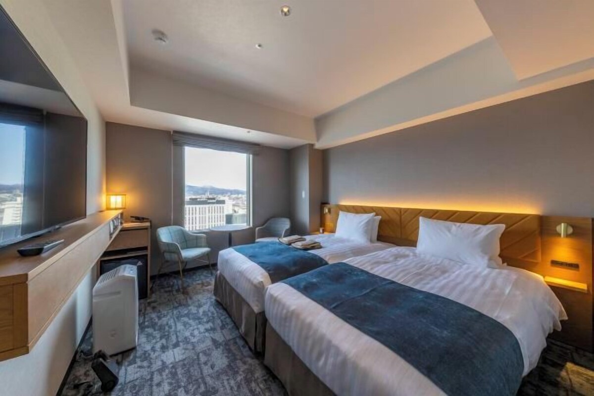 A hotel room with two beds, a desk, a chair, a TV, and a window with a city view.