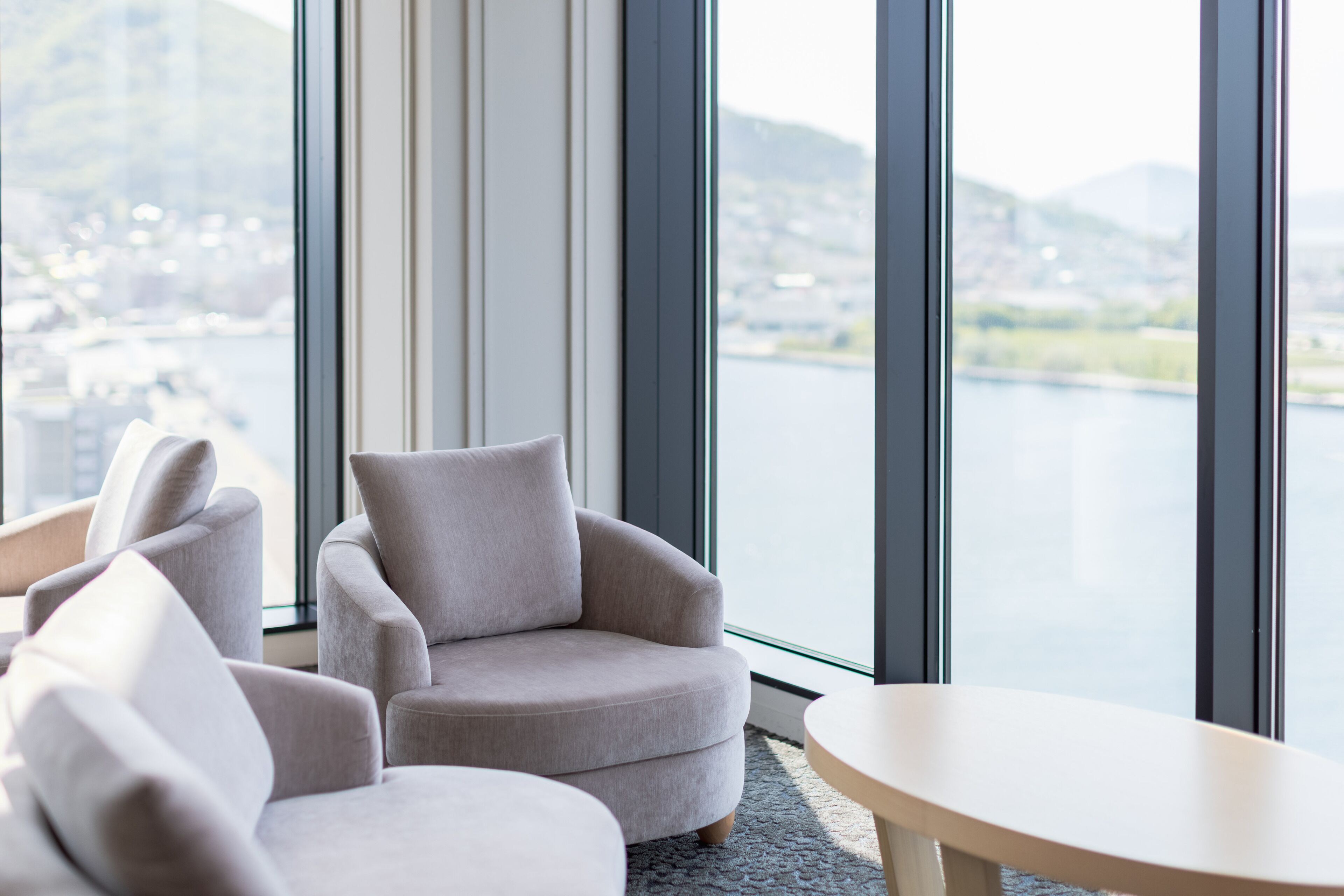Une chambre moderne dotée d’une grande fenêtre donnant sur un lac et sur des bâtiments situés au loin. On y trouve deux fauteuils gris et une petite table ronde.