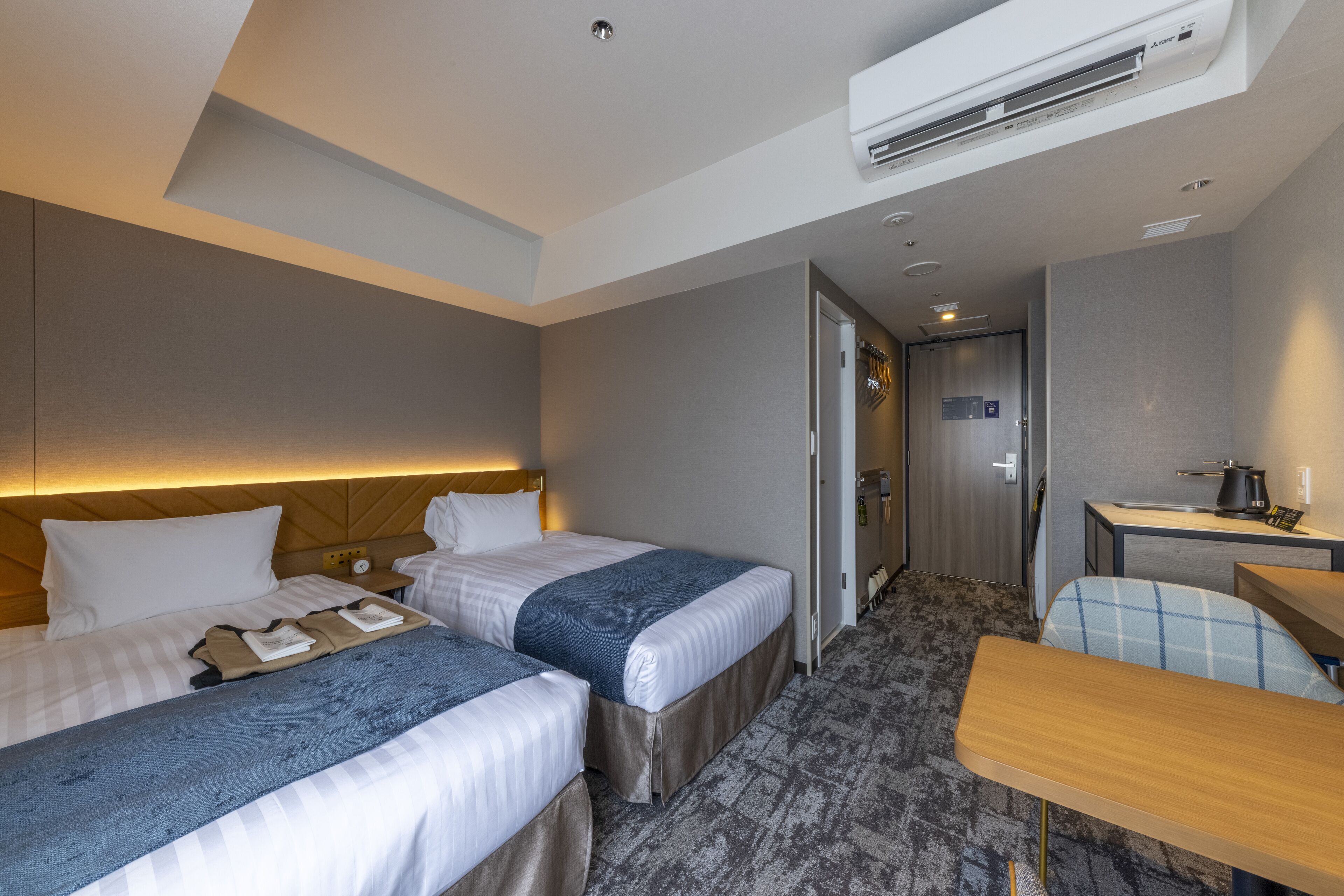 A hotel room with two beds, a desk, a chair, and a kitchenette.