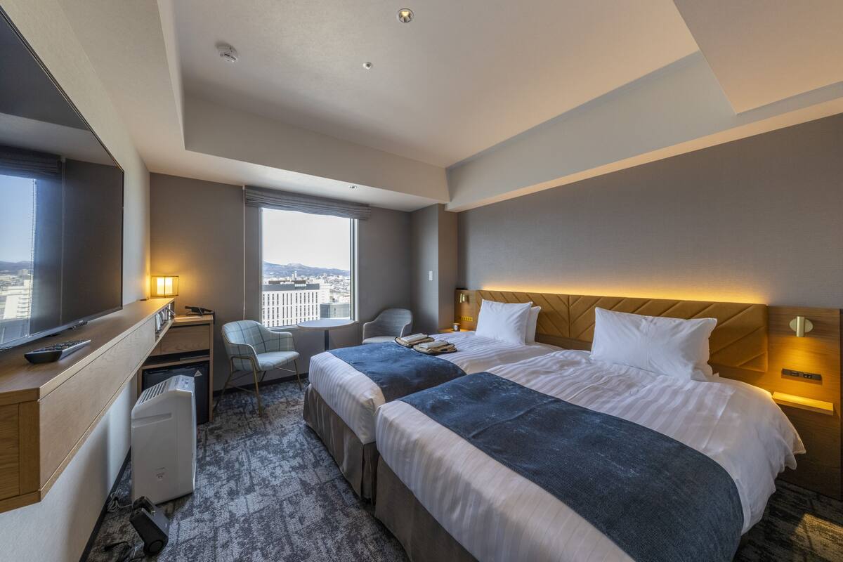 Habitación de hotel con dos camas, un escritorio, una silla, un televisor y una ventana con vistas a la ciudad.
