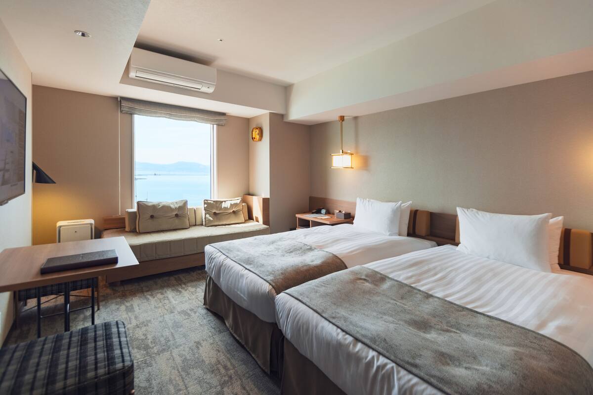 Habitación de hotel con dos camas, un escritorio, una silla, un ventanal con vista al mar y un reloj en la pared.