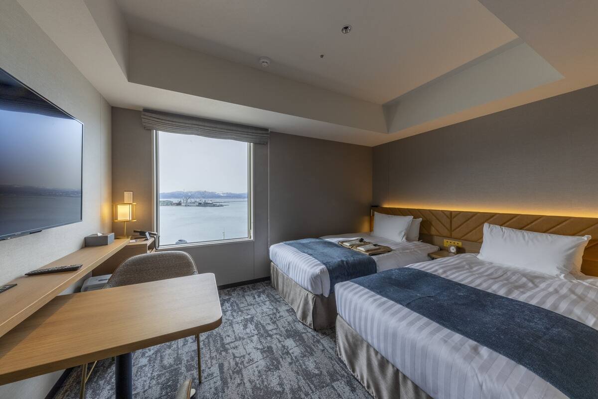 Habitación de hotel con dos camas, un escritorio, un televisor y una ventana con vistas al agua.