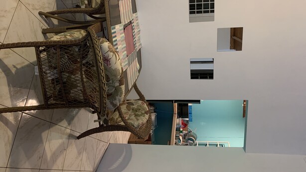 Quarto com piso de azulejos, uma cadeira de vime, uma mesinha e vista para uma piscina pela janela.