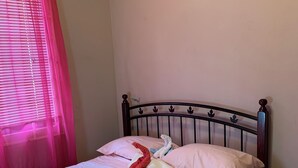 Uma cama com colcha rosa e branca, uma mesinha de cabeceira de madeira e uma janela com cortinas rosa.
