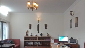 Una sala de estar con un sofá rojo, una mesa de centro de madera y un escritorio con una computadora. Se observa una estantería con objetos decorativos y una araña.