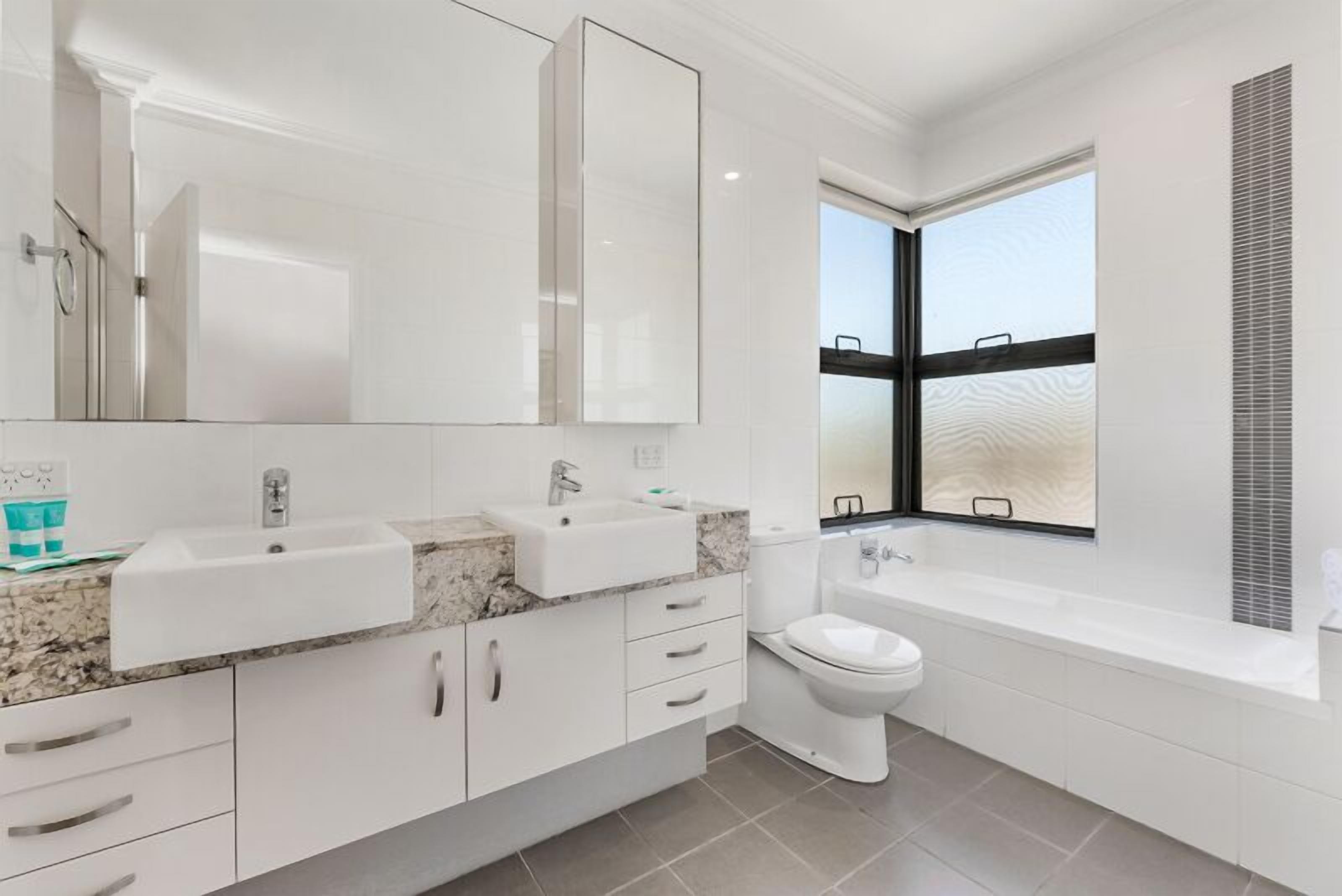 A modern bathroom with a large mirror, two sinks, a toilet, and a bathtub.