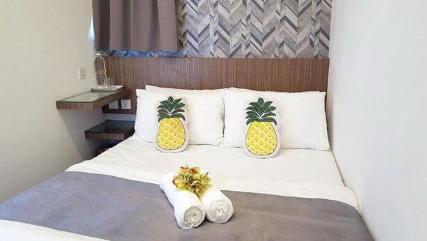 A neatly made bed with white bedding and two pineapple-shaped pillows, a small bedside table with a glass container, and a wall with a herringbone pattern.