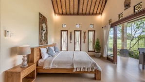 A bedroom with a wooden bed, bedside tables, a lamp, and a view of the outdoors through large windows. - Villa Kubu Agung (Ubud)