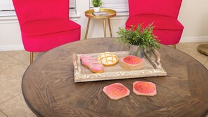 A round wooden table with a tray holding pink and white cookies, a potted plant, and a magazine. Two bright red chairs are positioned behind the table. - Fun Downtown Home Near Riverwalk (San Antonio)