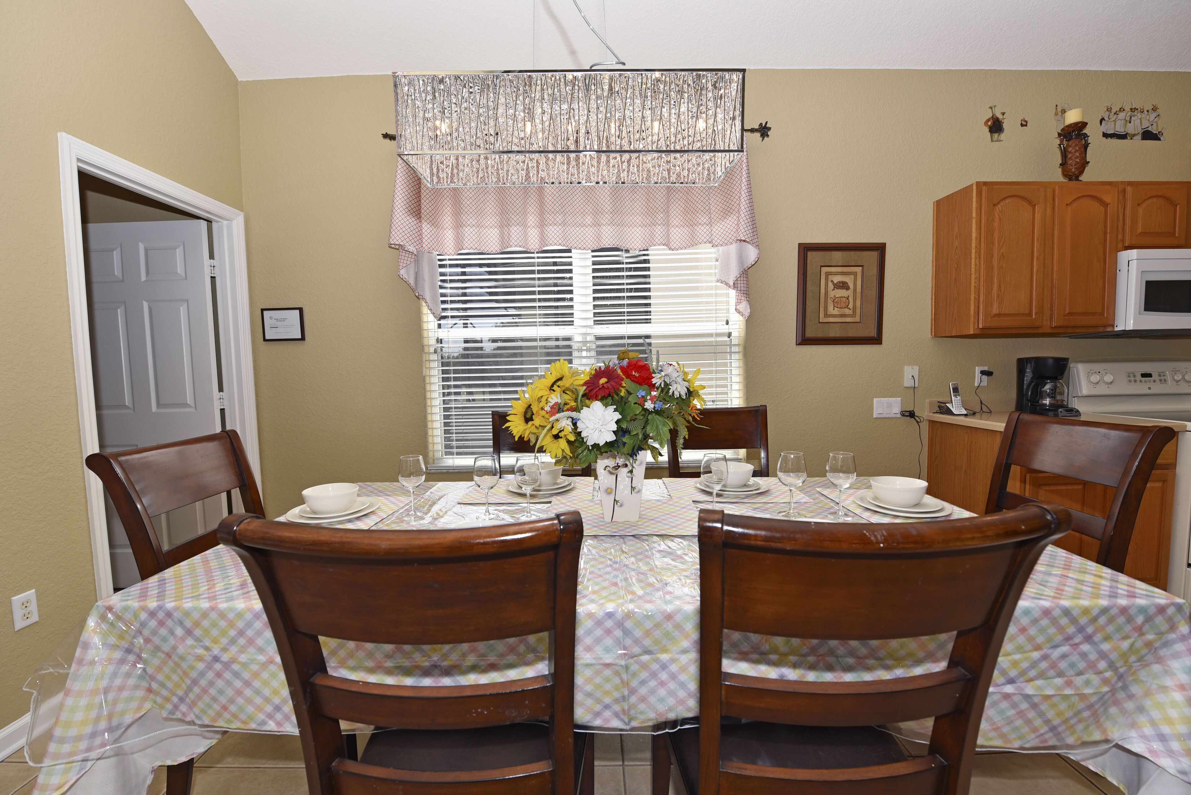 A dining room with a table set for six, wooden chairs, a vase with flowers, and a window with blinds.