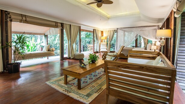 A spacious living room with a wooden floor, a ceiling fan, a sofa, a coffee table, and a view of greenery outside. - Galalima Glamping (Selemadeg)