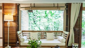 A wooden sofa with cushions, a wooden coffee table, a patterned rug, a floor lamp, and a view of greenery through a window. - Galalima Glamping (Selemadeg)