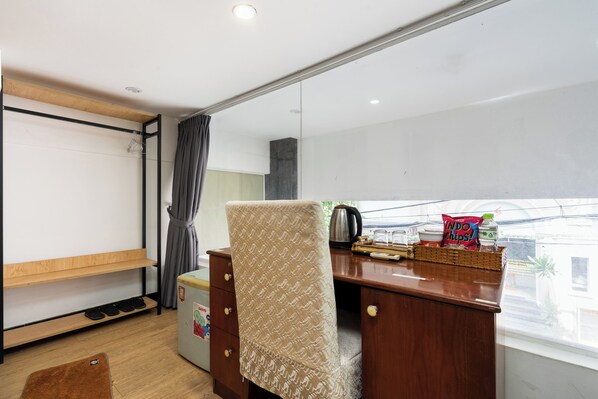 Quarto com escrivaninha de madeira, uma cadeira, pequeno frigorífico e janela com vista para edifícios.