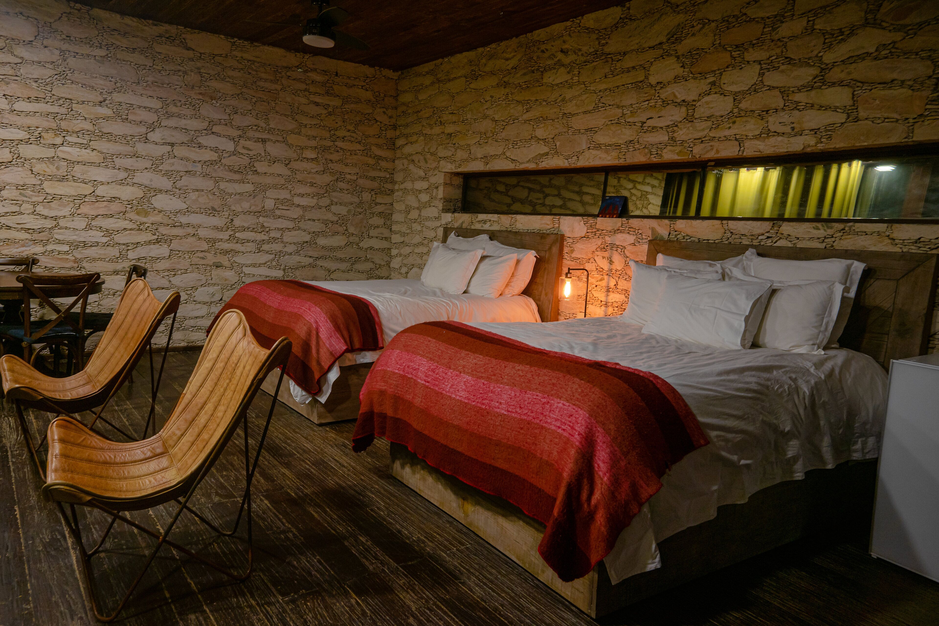 A room with a stone wall, a bed with white bedding, two wooden chairs, and a red blanket on the bed.