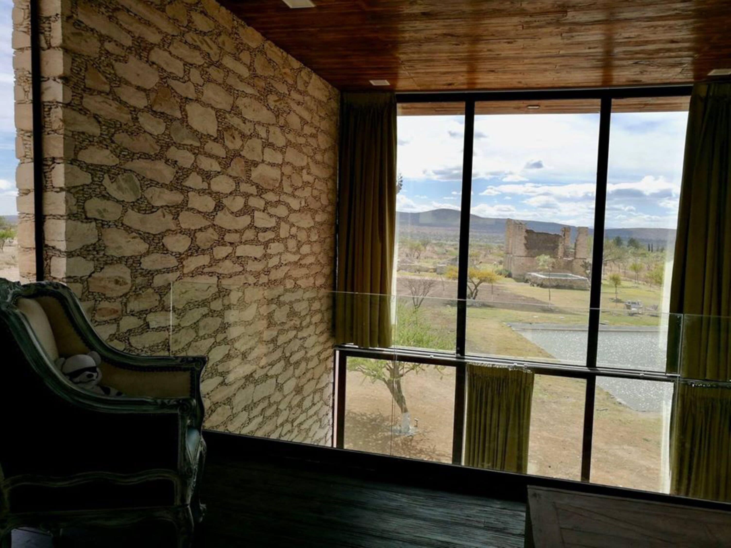 A room with a stone wall, a large window with curtains, and a view of a landscape including a building and a river.