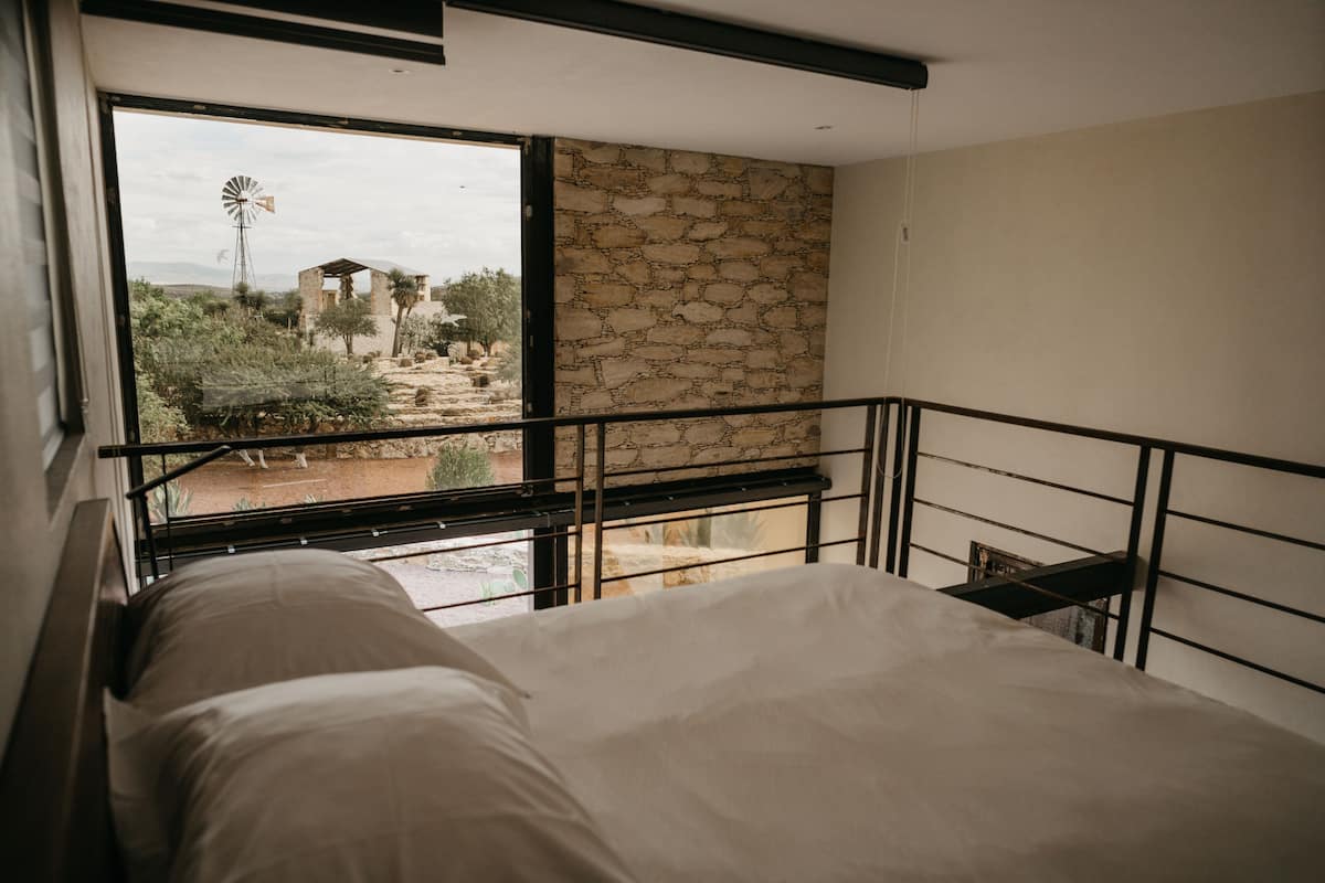 A bedroom with a large window, a stone wall, and a view of a windmill and landscape.