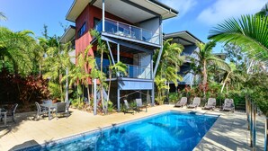 Una casa moderna de dos pisos con piscina, mobiliario exterior y plantas tropicales.