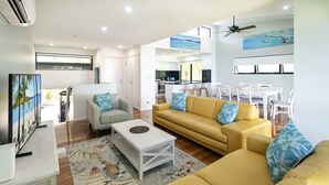 Una sala de estar moderna con un sofá amarillo, una mesa de centro blanca y un tapete estampado. Hay una zona de cocina con armarios blancos y una mesa de comedor con sillas.
