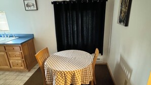 Een gezellig eetgedeelte met een ronde tafel, bedekt met een geruit tafelkleed, twee houten stoelen, een raam met donkere gordijnen, een keuken met een spoelbak en kastjes, en een aan de wand hangend schilderij.