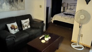 Een hotelkamer met een zwarte leren bank, een bijzettafeltje met decoratieve objecten, een bed met een zwart hoofdeinde en een staande ventilator.