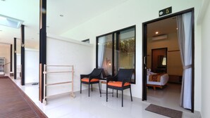A modern hotel room with a large sliding glass door leading to a balcony, two black chairs with orange cushions, a wooden ladder, and a bed with a white duvet and a brown throw blanket.
